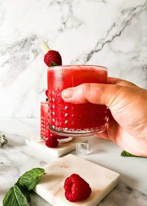 hand holding an apple cider vinegar mocktail, one more mocktail behind, raspberries , mint and ice around, on marble coasters and on a marble background