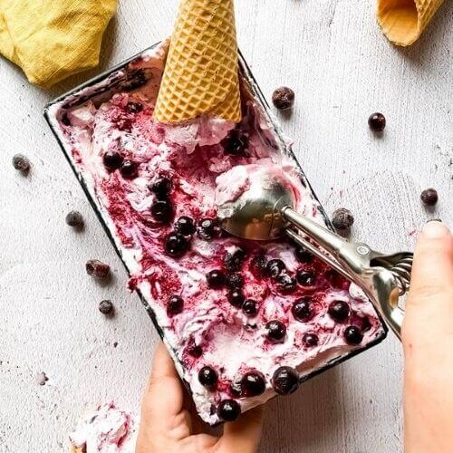 Blackcurrant Ice Cream: The Delicious Summer Recipe 6 blackcurrant ice cream in a tin with ice cream cones around, hands holding an ice cream scooper, blackcurrants around