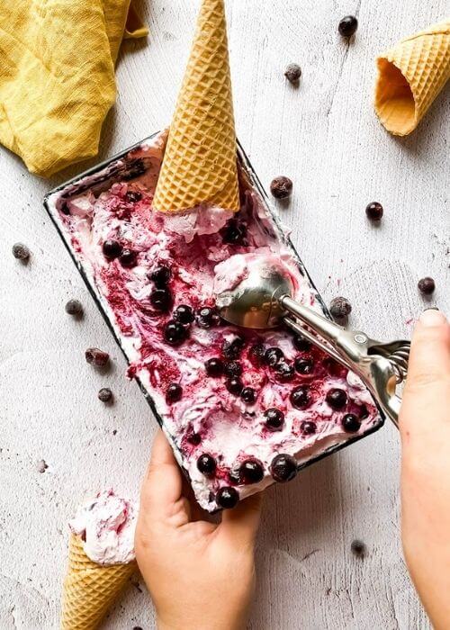 blackcurrant ice cream in a tin with ice cream cones around, hands holding an ice cream scooper, blackcurrants around
