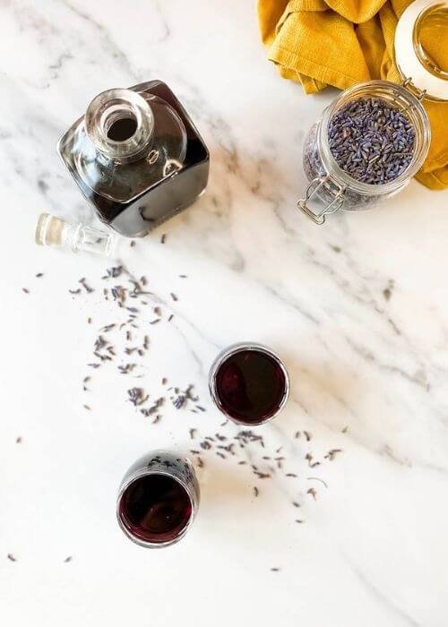 Lavender Liqueur Recipe - How To Make Delicious Lavande Liqueurs 4 from top - lavender liqueur in a bottle with two glasses, lavender buds in a jar and around, yellow towel on the side, on a marble background