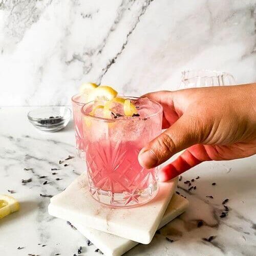 Delicious Lavender Mocktail, Easy To Make 6 lavender mocktail in a glass, a hand holding it in front, lavender buds, lemon slices and jars around on marble background