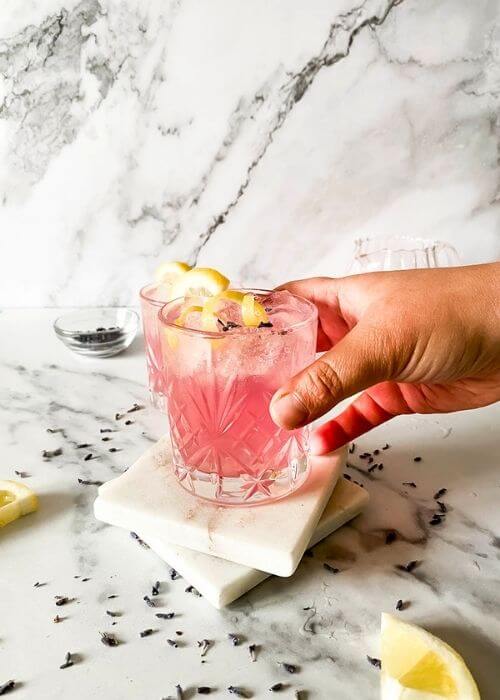 lavender mocktail in a glass, a hand holding it in front, lavender buds, lemon slices and jars around on marble background