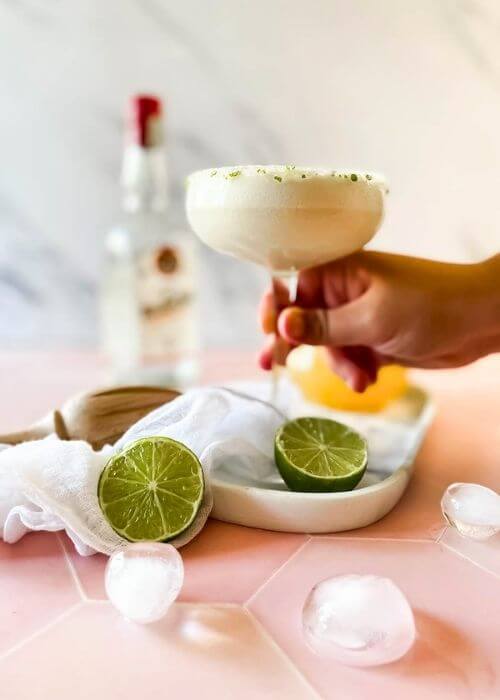 The #1 Best Pineapple Coconut Margarita Recipe 2 from front - pineapple coconut margarita in a glass, held by a hand, on a marble tray, limes, ice cubes around, in the back a lemon squeezer and a bottle, on marble and pink background