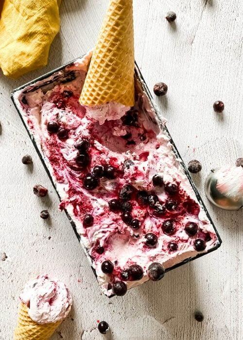 Blackcurrant Ice Cream: The Delicious Summer Recipe 5 blackcurrant ice cream in a tin with ice cream cones around, an ice cream scooper, blackcurrants around