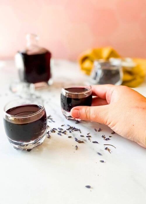 Lavender Liqueur Recipe - How To Make Delicious Lavande Liqueurs 2 hand holding one of the glasses, lavender liqueur in a bottle with two glasses, lavender buds in a jar and around, yellow towel on the side, on a marble background