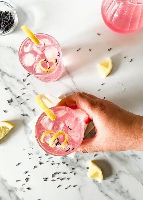 Delicious Lavender Mocktail, Easy To Make 5 from top - two lavender mocktail in a glasses, a hand holding one, lavender buds, lemon slices and jars around on marble background