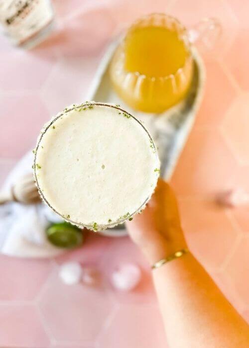 The #1 Best Pineapple Coconut Margarita Recipe 3 from top -hand holding a pineapple margarita in a glass on a marble tray, limes, ice cubes around, in the back a lemon squeezer and a bottle, on marble and pink background