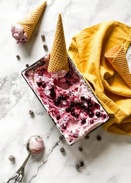 Blackcurrant Ice Cream: The Delicious Summer Recipe 4 from top - blackcurrant ice cream in a tin with ice cream cones around, an ice cream scooper, blackcurrants around