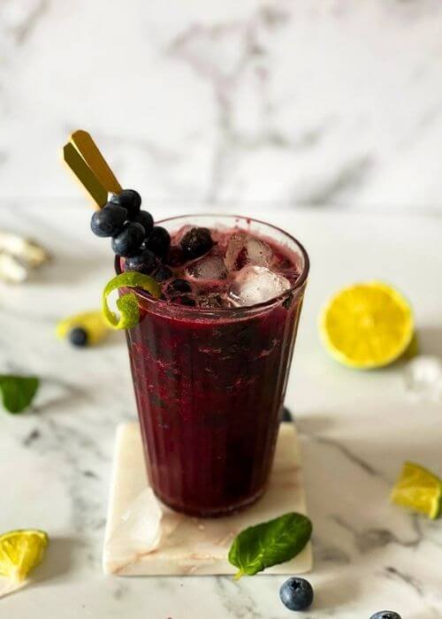 Delicious Blueberry Mojito Mocktail Recipe 3 from front -.blueberry mojito mocktail in a glass with ingredients around on marble coaster and marble background