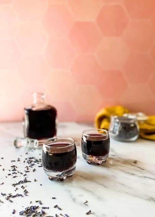 Lavender Liqueur Recipe - How To Make Delicious Lavande Liqueurs 3 from front - lavender liqueur in a bottle with two glasses, lavender buds in a jar and around, yellow towel on the side, on a marble background