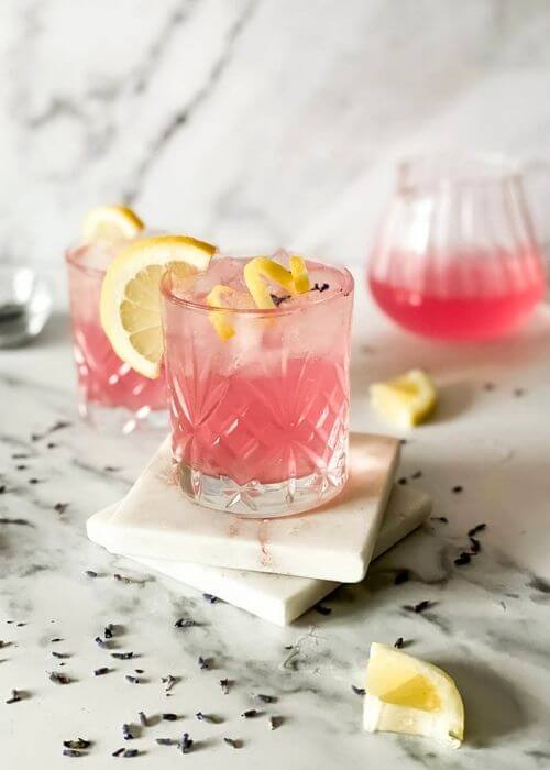 Delicious Lavender Mocktail, Easy To Make 3 two lavender mocktail in a glasses, lavender buds, lemon slices and jars around on marble background