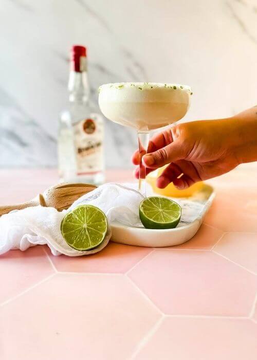 The #1 Best Pineapple Coconut Margarita Recipe 5 from front - pineapple coconut margarita, hand holding it, in a glass on a marble tray, limes, ice cubes around, in the back a lemon squeezer and a bottle, on marble and pink background