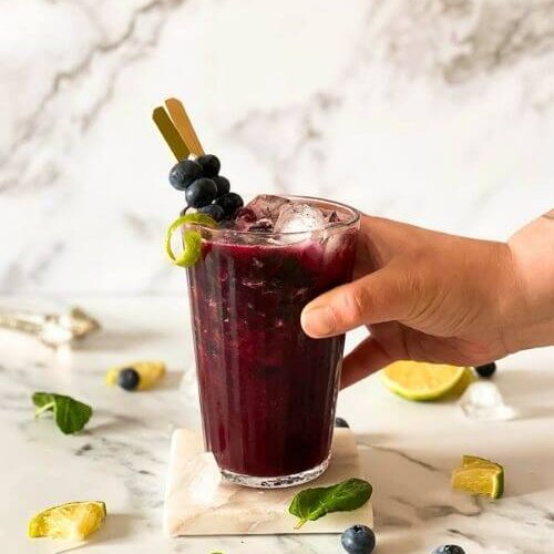 Delicious Blueberry Mojito Mocktail Recipe 5 hand holding a blueberry mocktail in a glass, ingredients around on marble background