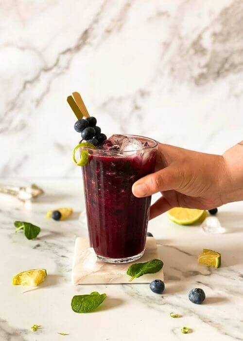 hand holding a blueberry mocktail in a glass, ingredients around on marble background
