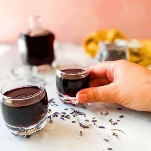 Lavender Liqueur Recipe - How To Make Delicious Lavande Liqueurs 5 hand holding a glass of liqueur, bottle with two glasses, lavender buds in a jar and around, yellow towel on the side, on a marble background