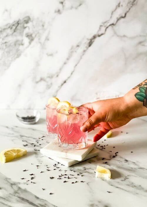 Delicious Lavender Mocktail, Easy To Make 4 two mocktail in a glasses, a hand holding one in front, lavender buds, lemon slices and jars around on marble background