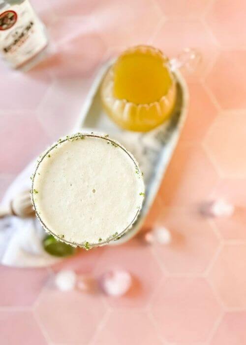 The #1 Best Pineapple Coconut Margarita Recipe 4 from top - pineapple coconut margarita in a glass on a marble tray, limes, ice cubes around, in the back a lemon squeezer and a bottle, on marble and pink background