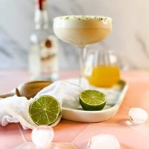 The #1 Best Pineapple Coconut Margarita Recipe 6 from front - pineapple coconut margarita in a glass on a marble tray, limes, ice cubes around, in the back a lemon squeezer and a bottle, on marble and pink background