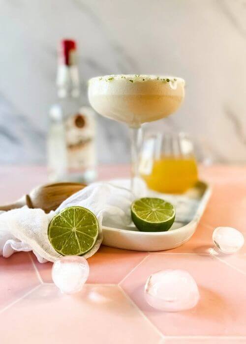from front - pineapple coconut margarita in a glass on a marble tray, limes, ice cubes around, in the back a lemon squeezer and a bottle, on marble and pink background
